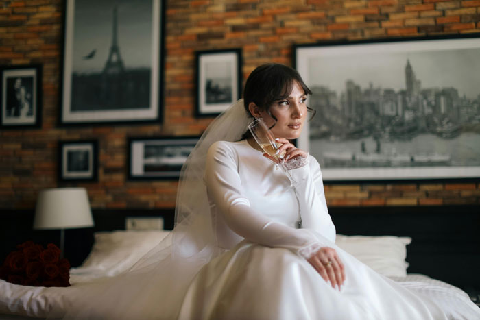 Bride in a white dress and veil, sitting on a bed, holding a champagne glass, in a room with framed pictures on the wall. Bride in a white dress and veil, sitting on a bed, holding a champagne glass, in a room with framed pictures on the wall.