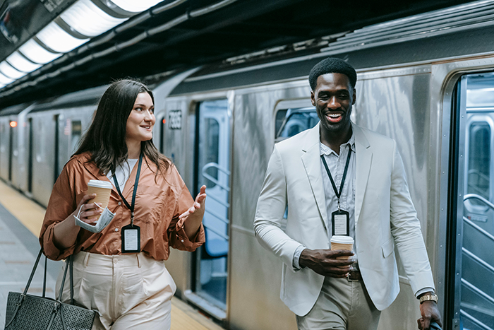 A man and woman in business attire walking in a subway station, holding coffee and talking, both wearing ID badges. A man and woman in business attire walking in a subway station, holding coffee and talking, both wearing ID badges.