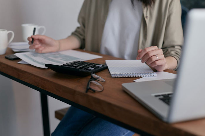 Person working with papers, calculator, and laptop on a wooden desk, planning a strategy for revenge. Person working with papers, calculator, and laptop on a wooden desk, planning a strategy for revenge.