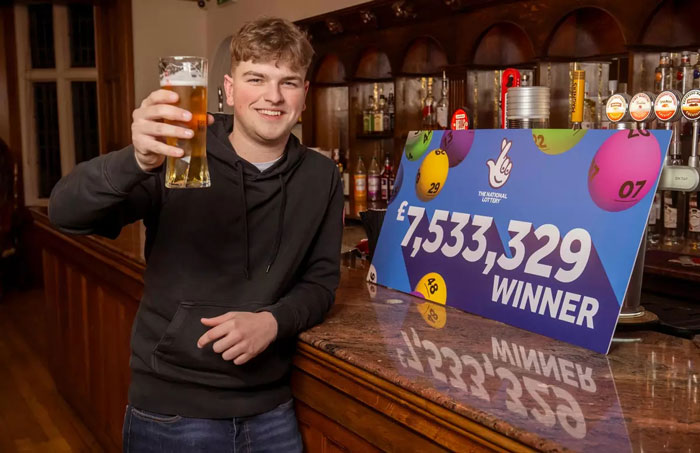 Man in a pub celebrating a £7.5M lottery win with a pint, standing next to a winning sign. Man in a pub celebrating a £7.5M lottery win with a pint, standing next to a winning sign.