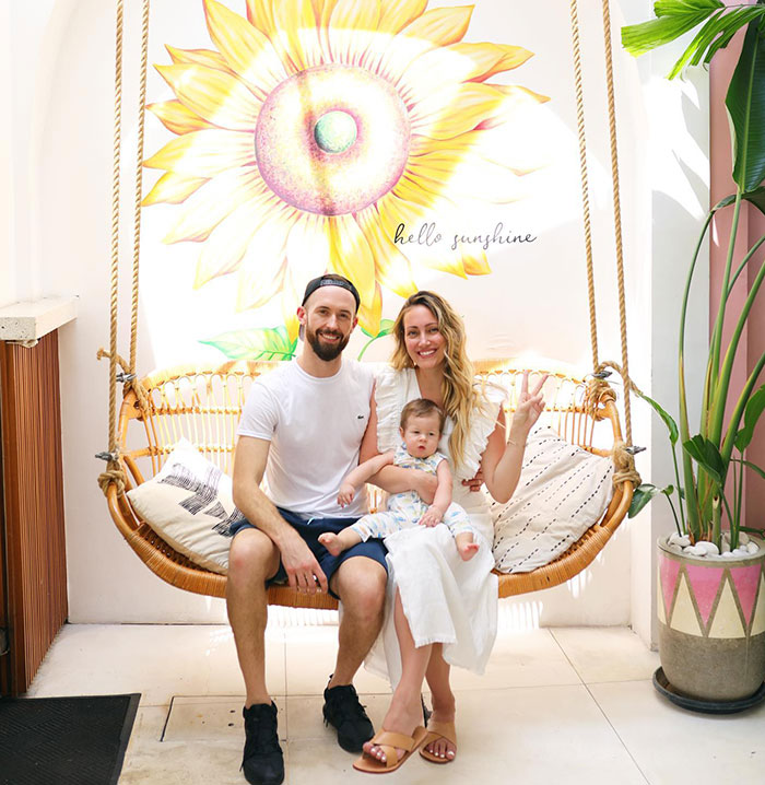 Family sitting on a swing, smiling, with a sunflower mural and the text "hello sunshine" in the background. Family sitting on a swing, smiling, with a sunflower mural and the text "hello sunshine" in the background.