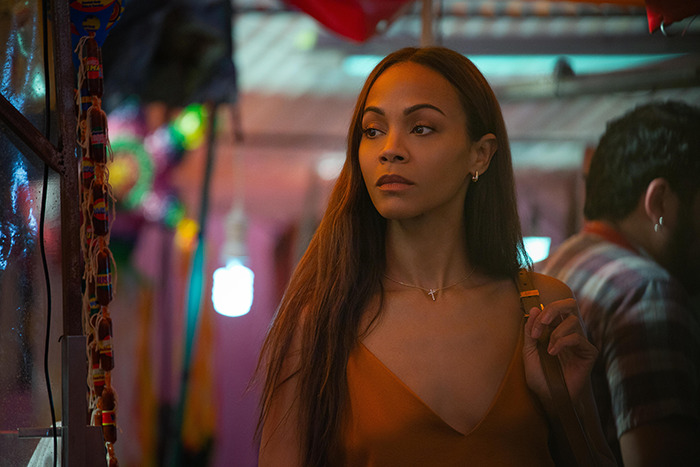 A woman in an orange top stands in a colorful market setting, looking contemplative. A woman in an orange top stands in a colorful market setting, looking contemplative.