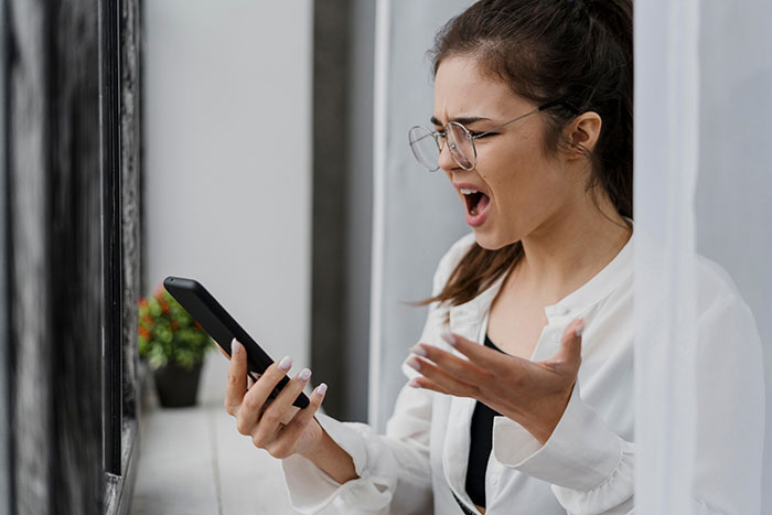 A woman in glasses looks frustrated while holding a phone, connected to an influencer controversy over free painting demands. A woman in glasses looks frustrated while holding a phone, connected to an influencer controversy over free painting demands.
