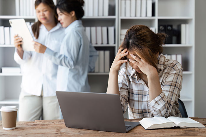 A person stressed at a desk with laptop, while two managers chat in the background, creating an uncomfortable atmosphere. A person stressed at a desk with laptop, while two managers chat in the background, creating an uncomfortable atmosphere.
