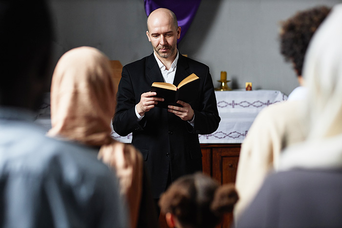 Religious leader reads to congregation, holding a book, standing in front of an altar, people listening attentively. Religious leader reads to congregation, holding a book, standing in front of an altar, people listening attentively.