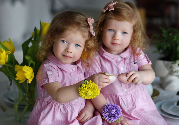 Twins in pink dresses holding flowers and eggs, highlighting different outfits for individuality. Twins in pink dresses holding flowers and eggs, highlighting different outfits for individuality.