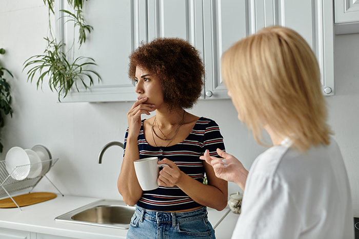 Two women in a kitchen, one holding a mug, discussing the old timers way of testing a woman. Two women in a kitchen, one holding a mug, discussing the old timers way of testing a woman.