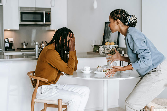 Couple having an argument at the kitchen table; man looks tired, covering his face, while woman gestures expressively. Couple having an argument at the kitchen table; man looks tired, covering his face, while woman gestures expressively.