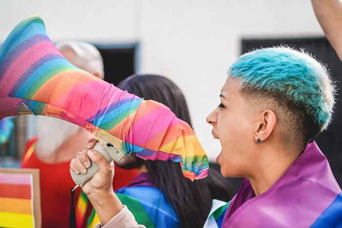 Person with blue hair and rainbow flag shouting through a megaphone covered in a rainbow cloth, highlighting hate crime awareness. Person with blue hair and rainbow flag shouting through a megaphone covered in a rainbow cloth, highlighting hate crime awareness.