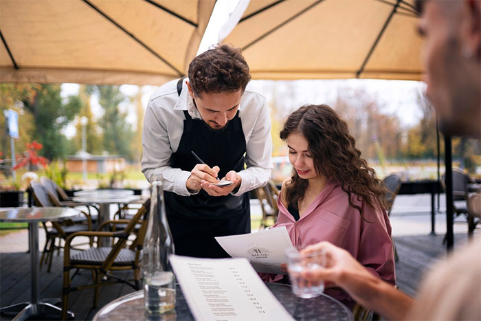 A bartender taking an order from a woman under a patio umbrella at an outdoor cafe. A bartender taking an order from a woman under a patio umbrella at an outdoor cafe.