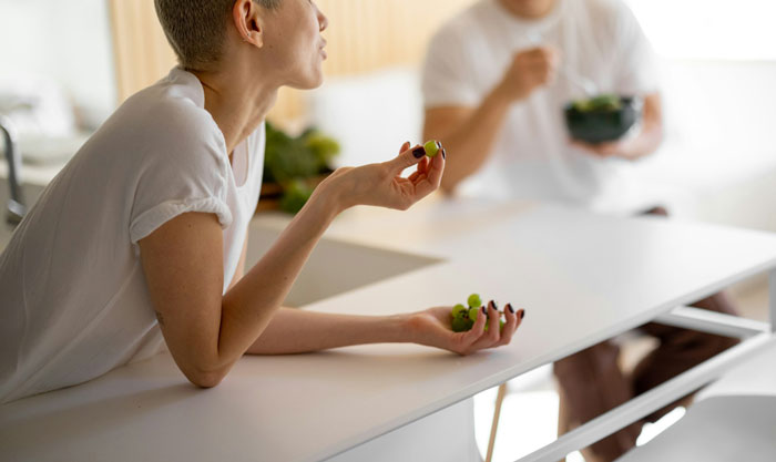 Teen in a white shirt talking, holding a grape, seated at a table in a bright kitchen setting. Teen in a white shirt talking, holding a grape, seated at a table in a bright kitchen setting.
