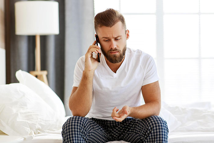 Man on phone looking concerned, sitting on bed, wearing checkered pants; a lamp is visible in the background. Man on phone looking concerned, sitting on bed, wearing checkered pants; a lamp is visible in the background.