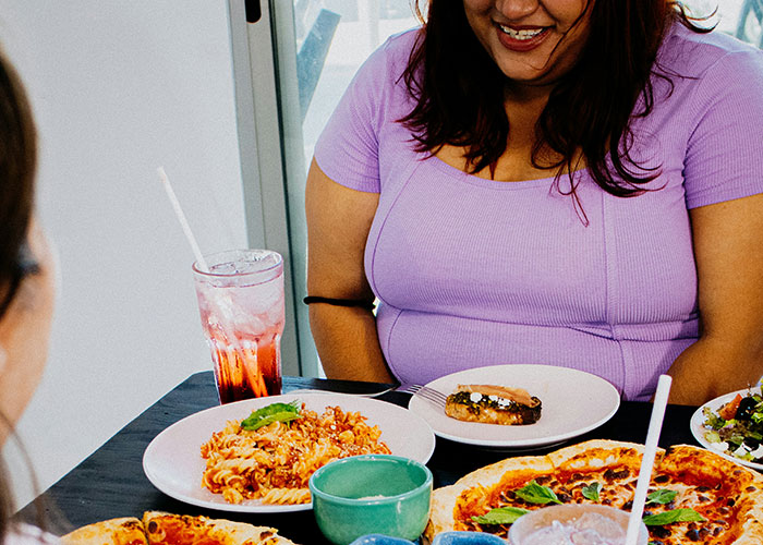 A woman in a purple top sitting at a table with pizza and pasta, reflecting on being overweight.