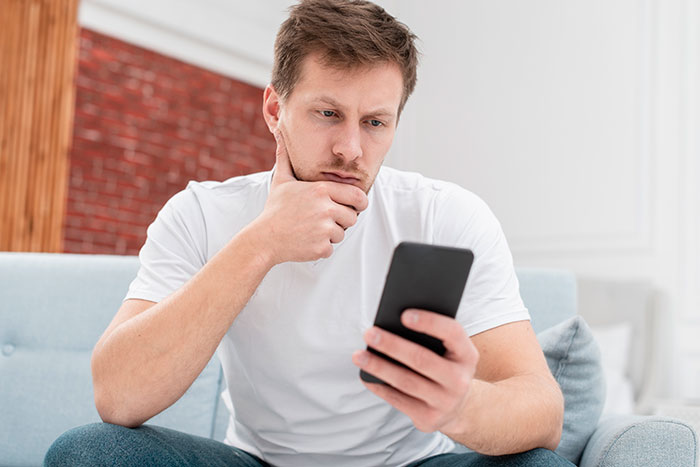 Man in white shirt on couch looking at phone, pondering family responsibilities. Man in white shirt on couch looking at phone, pondering family responsibilities.