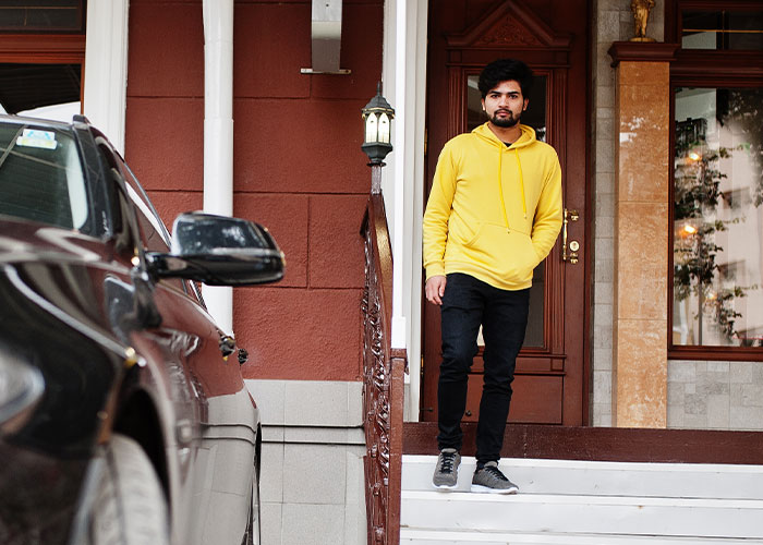 Man in a yellow hoodie on a porch looks at a car parked in a neighbor's driveway. Man in a yellow hoodie on a porch looks at a car parked in a neighbor's driveway.