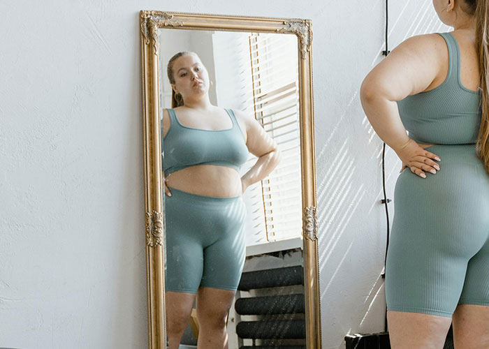 Person in blue activewear looking at their reflection in a full-length mirror, highlighting aspects of being overweight.
