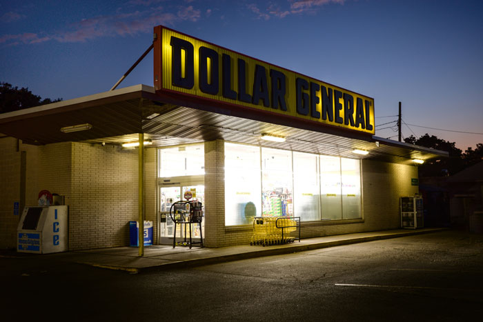 Dollar General store at dusk, illuminated with bright fluorescent lights, highlighting contrast with wealthy lifestyles.