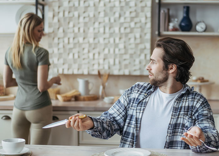 Man at a kitchen table with a knife, looking towards a woman; concept of husband’s ungrateful birthday meltdown. Man at a kitchen table with a knife, looking towards a woman; concept of husband’s ungrateful birthday meltdown.