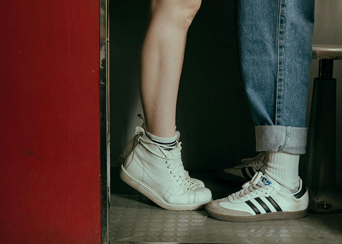 Two people in casual sneakers standing next to each other, one in white high-tops, the other in Adidas, indoors.