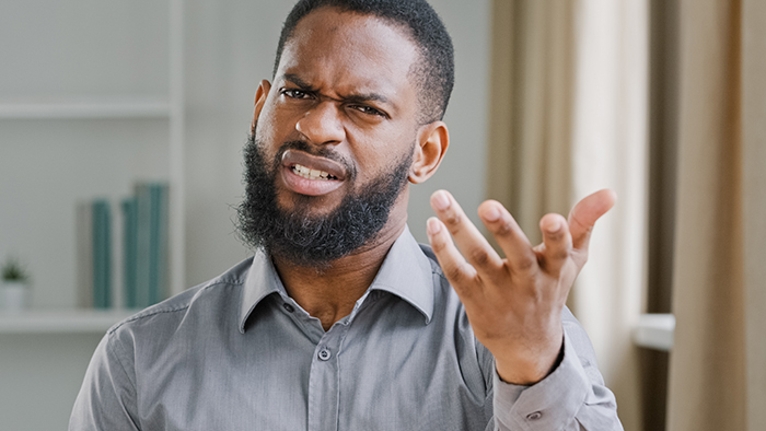Man in a gray shirt looking puzzled, hand raised, in a room with curtains, related to office outfit discussion. Man in a gray shirt looking puzzled, hand raised, in a room with curtains, related to office outfit discussion.