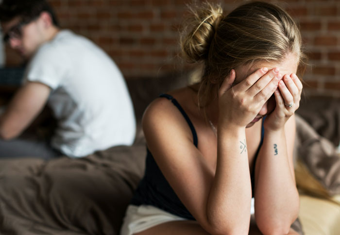 Woman with hands on face, appearing upset, while man sits on bed, facing away. Woman with hands on face, appearing upset, while man sits on bed, facing away.
