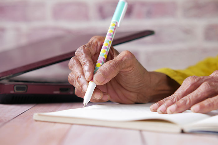Older woman writing with a colorful pen near a laptop, contemplating inheritance decisions. Older woman writing with a colorful pen near a laptop, contemplating inheritance decisions.