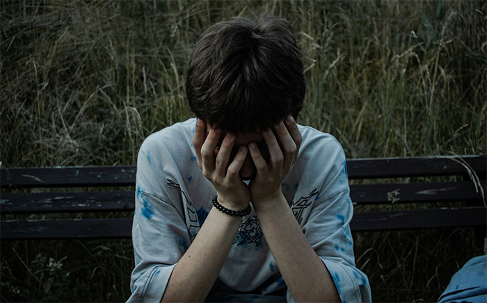 Teen sitting on a bench with head in hands, wearing a patterned shirt, in a natural outdoor setting. Teen sitting on a bench with head in hands, wearing a patterned shirt, in a natural outdoor setting.