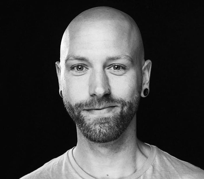 Bald man with a beard wearing earrings, neutral expression, against a dark background, discussing Covid vaccines. Bald man with a beard wearing earrings, neutral expression, against a dark background, discussing Covid vaccines.