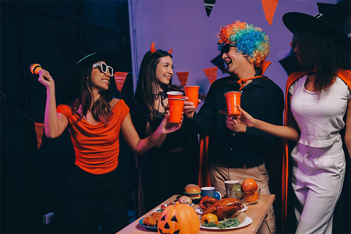 Friends in costume at a Halloween party, laughing and holding drinks, with a decorated table in the foreground. Friends in costume at a Halloween party, laughing and holding drinks, with a decorated table in the foreground.