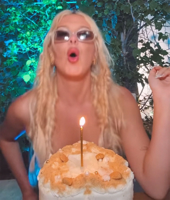 Blonde woman celebrating with a lit birthday candle on a cake, wearing sunglasses, greenery in the background. Blonde woman celebrating with a lit birthday candle on a cake, wearing sunglasses, greenery in the background.