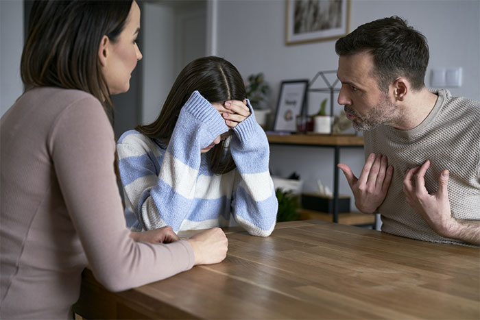 Teens arguing with parents at a table, one with head in hands, discussing sibling's sexuality and family beliefs. Teens arguing with parents at a table, one with head in hands, discussing sibling's sexuality and family beliefs.