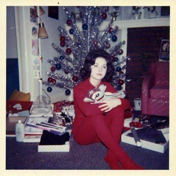 A vintage image of a woman in red by a retro Christmas tree, clutching a plush toy amidst gifts.