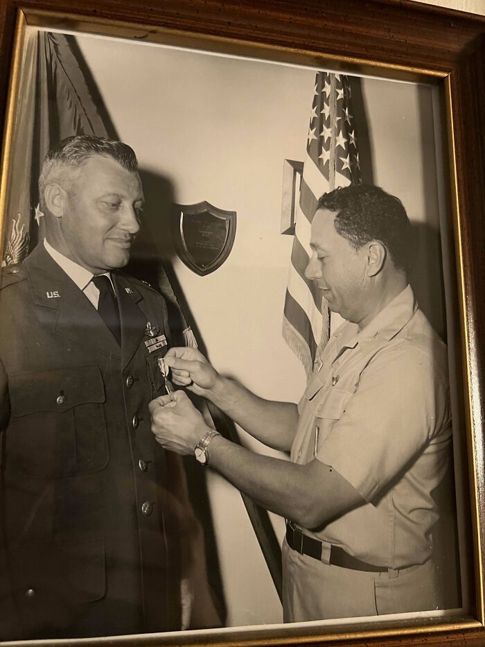 Historic image of two military officers, one pinning a medal on the other, with flags and a plaque in the background.
