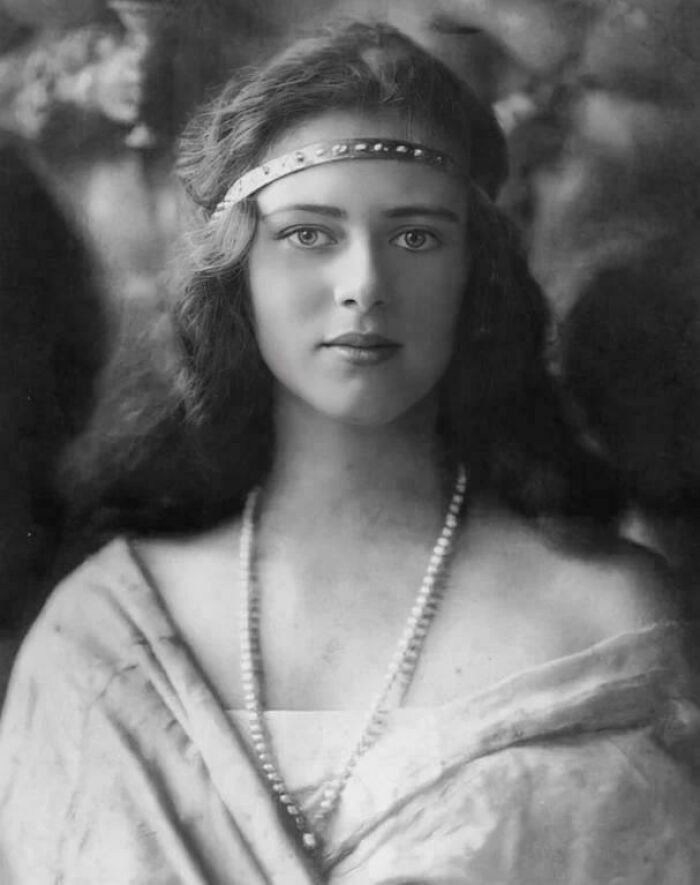 A historic portrait of a woman with long hair, wearing a headband and pearl necklace from the early 20th century.
