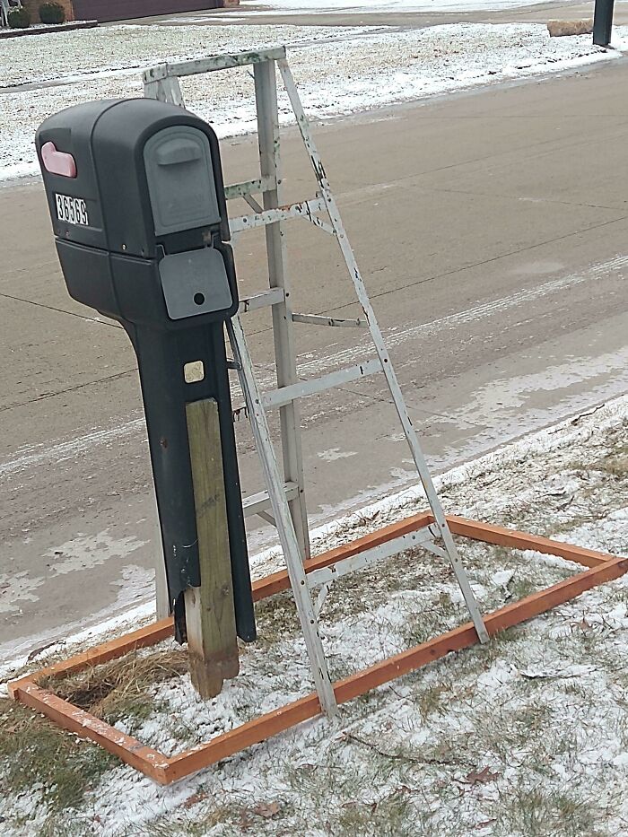 Mailbox supported by a wooden post with a metal ladder frame and a rectangular wooden base, showcasing redneck engineer creativity.