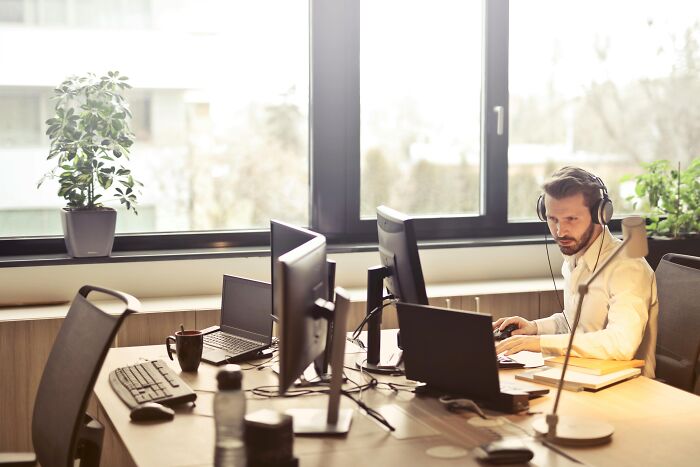 Man wearing headphones working at a desk with multiple monitors in an office setting, adding humor to the office environment.