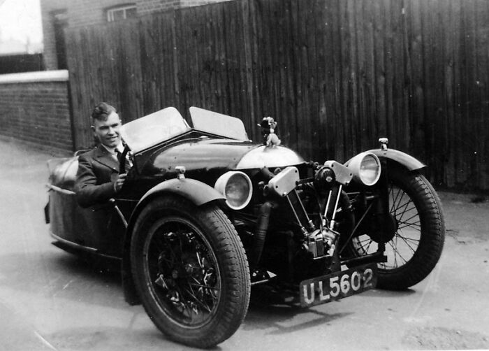 A man sitting in a vintage car parked on a street, showcasing early automotive design.