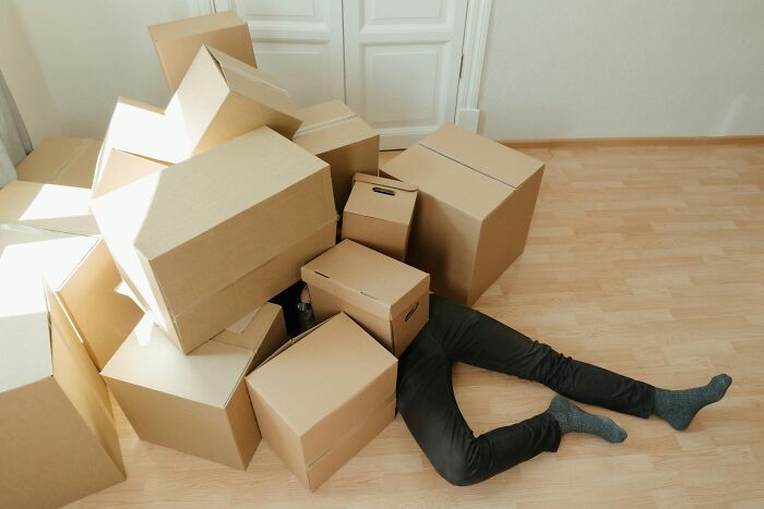 Person buried under a pile of cardboard boxes on a wooden floor, illustrating a humorous take on brilliant life hacks.