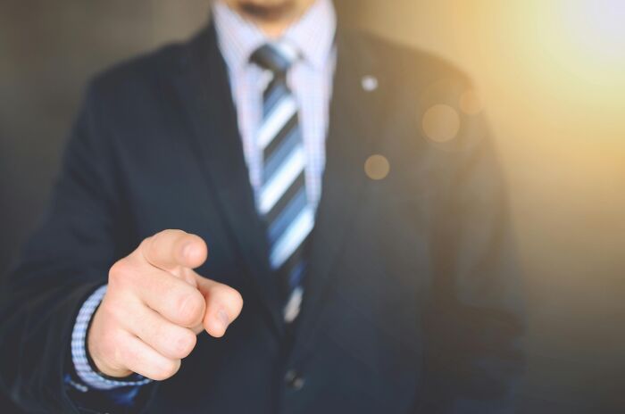 Business person pointing, symbolizing work-struggles in a formal setting, wearing a suit and tie with blurred background.