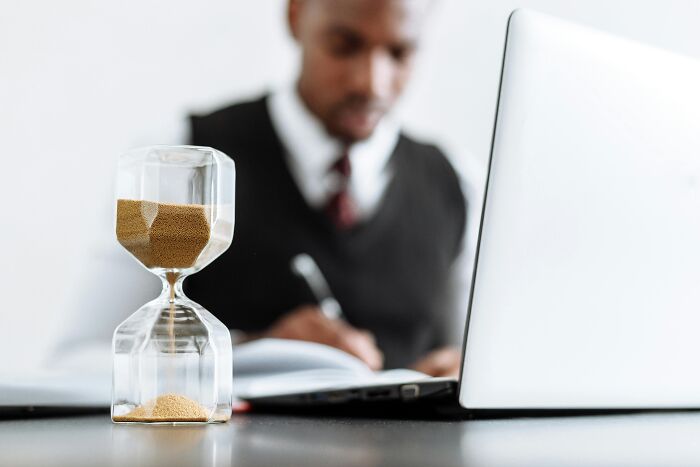 Hourglass on desk with man working on laptop, illustrating work-struggles and time management.