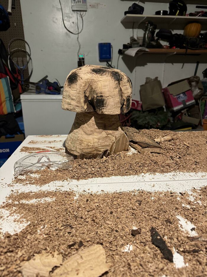 Woodworkers carving a mushroom-shaped project surrounded by wood shavings on a white workbench in a workshop.