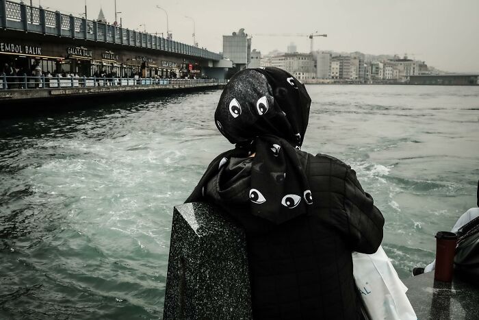 Person with eye-patterned headscarf gazes at a river under a cloudy sky, symbolizing a unique view of urban life.
