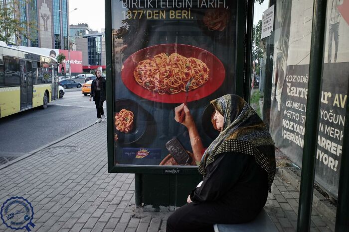 A woman sits at a bus stop with her hand perfectly aligned with a pasta poster, showcasing Gökhan Arer's unique street perspective.