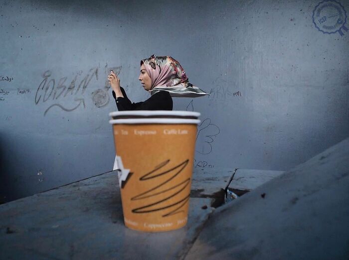 A woman with a headscarf appears behind a coffee cup, illustrating Gökhan Arer's unique street perspective.