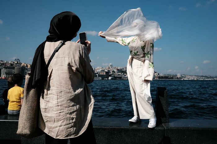 Person with a headscarf takes a photo near the water, capturing a breezy moment. Gökhan Arer's street photography.