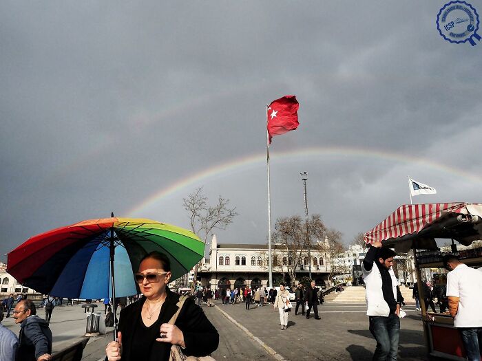 Woman with rainbow umbrella near flag and double rainbow; Gökhan Arer captures street life differently.