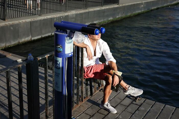 Man sitting by the water on a wooden boardwalk, inadvertently aligning his eyes with binoculars, creating a humorous street perspective.