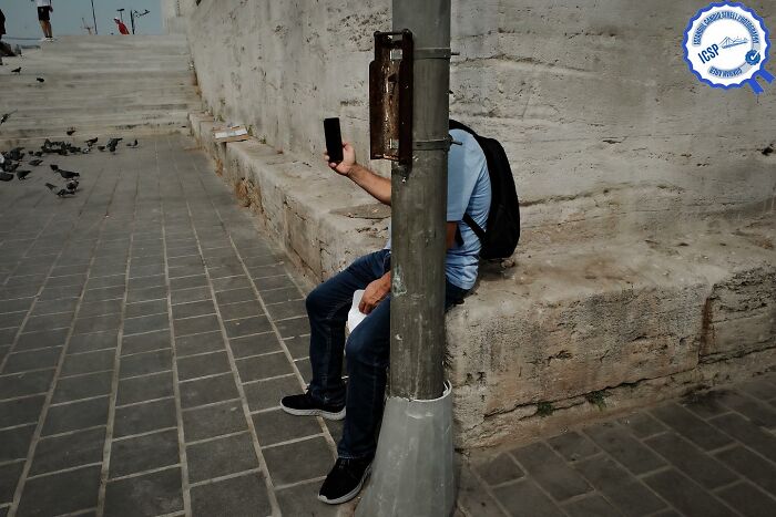 Street photography by Gökhan Arer showing a man perfectly obscured by a pole while taking a selfie.