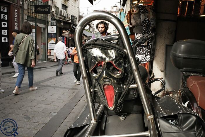 "Unique street scene with a mask on a motorcycle, people walking by, capturing Gökhan Arer's distinctive perspective."
