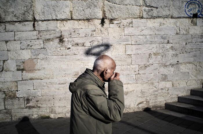 An elderly man in a coat walks against a stone wall with a shadow appearing like a halo, captured differently by Gökhan Arer.
